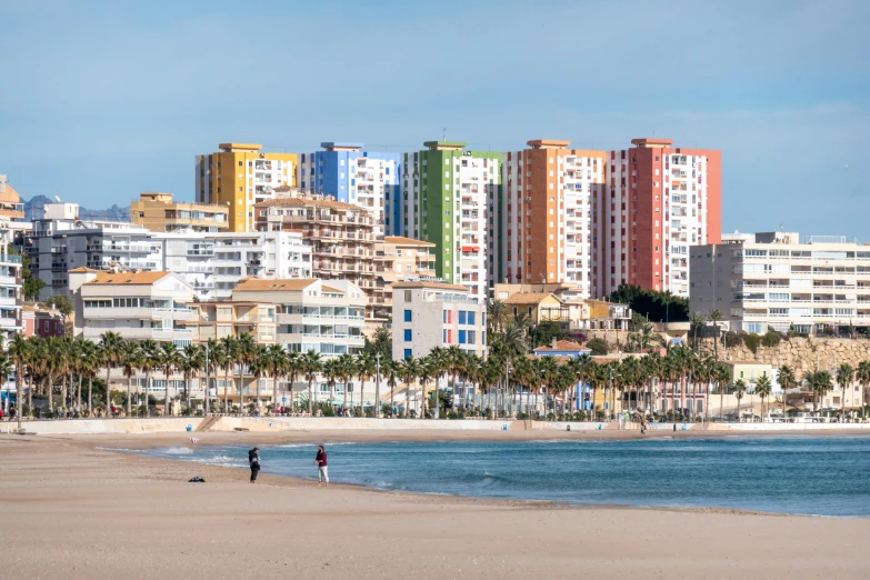 a couple of people walking on top of a sandy beach, a photo, by Carlo Martini, shutterstock, graffiti, brightly colored buildings, costa blanca, high rises, viewed from the ocean