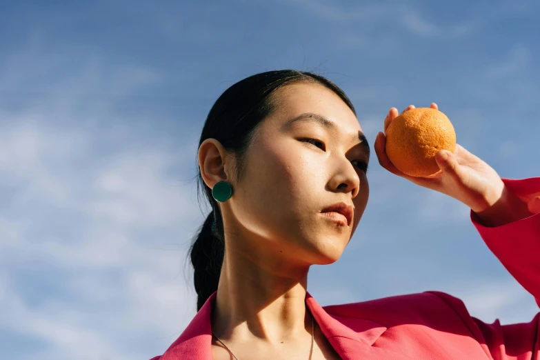 a woman holding an orange up to her face, inspired by Ren Hang, trending on pexels, ear, on a sunny day, ethnicity : japanese, round portruding chin