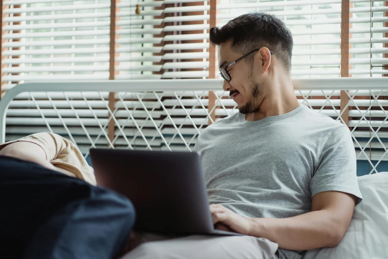 a man sitting on a bed using a laptop, trending on pexels, with nerdy glasses and goatee, two buddies sitting in a room, lachlan bailey, avatar image