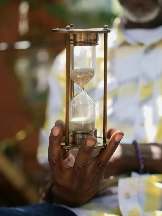 a man holding a hourglass in his hand, by Jessie Algie, unsplash, process art, african steampunk alchemist, instagram story, afternoon sunlight, community celebration