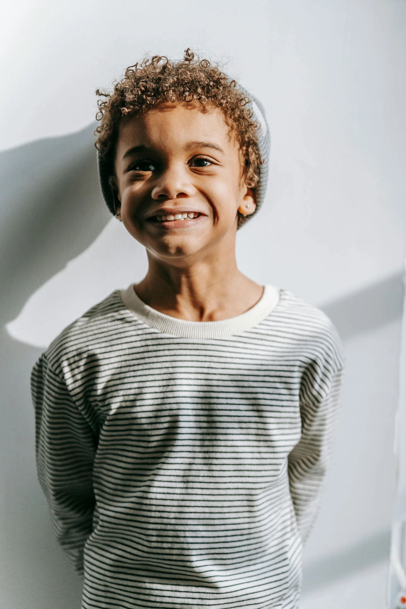 a young boy standing in front of a white wall, a character portrait, inspired by Louis Le Nain, pexels contest winner, wearing stripe shirt, large black smile, soft backlighting, long sleeve