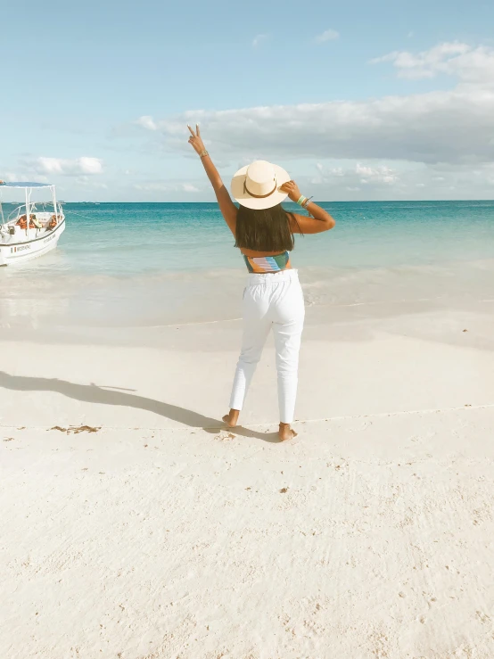 a woman standing on a beach next to a boat, white pants, carribean white sand, with arms up, instagram post
