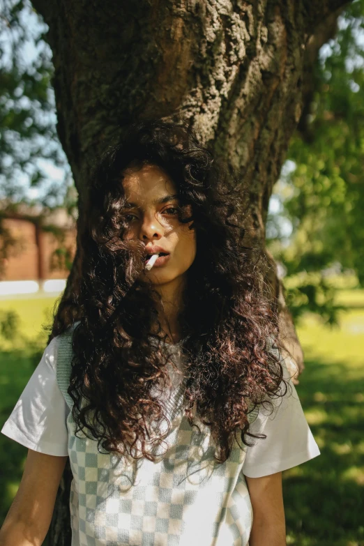 a woman standing next to a tree with a cigarette in her mouth, an album cover, inspired by Elsa Bleda, pexels contest winner, renaissance, black long curly hair, college, light skin, casual pose