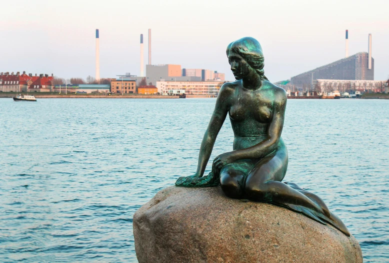 a statue of a mermaid sitting on a rock, a statue, by Jens Søndergaard, the sea seen behind the city, warm glow, harbour in background, bronze statue and silver