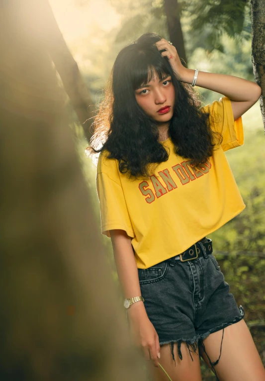 a woman standing next to a tree in a forest, inspired by Elsa Bleda, unsplash contest winner, graffiti, wearing a modern yellow tshirt, ulzzang, model エリサヘス s from acquamodels, curly bangs