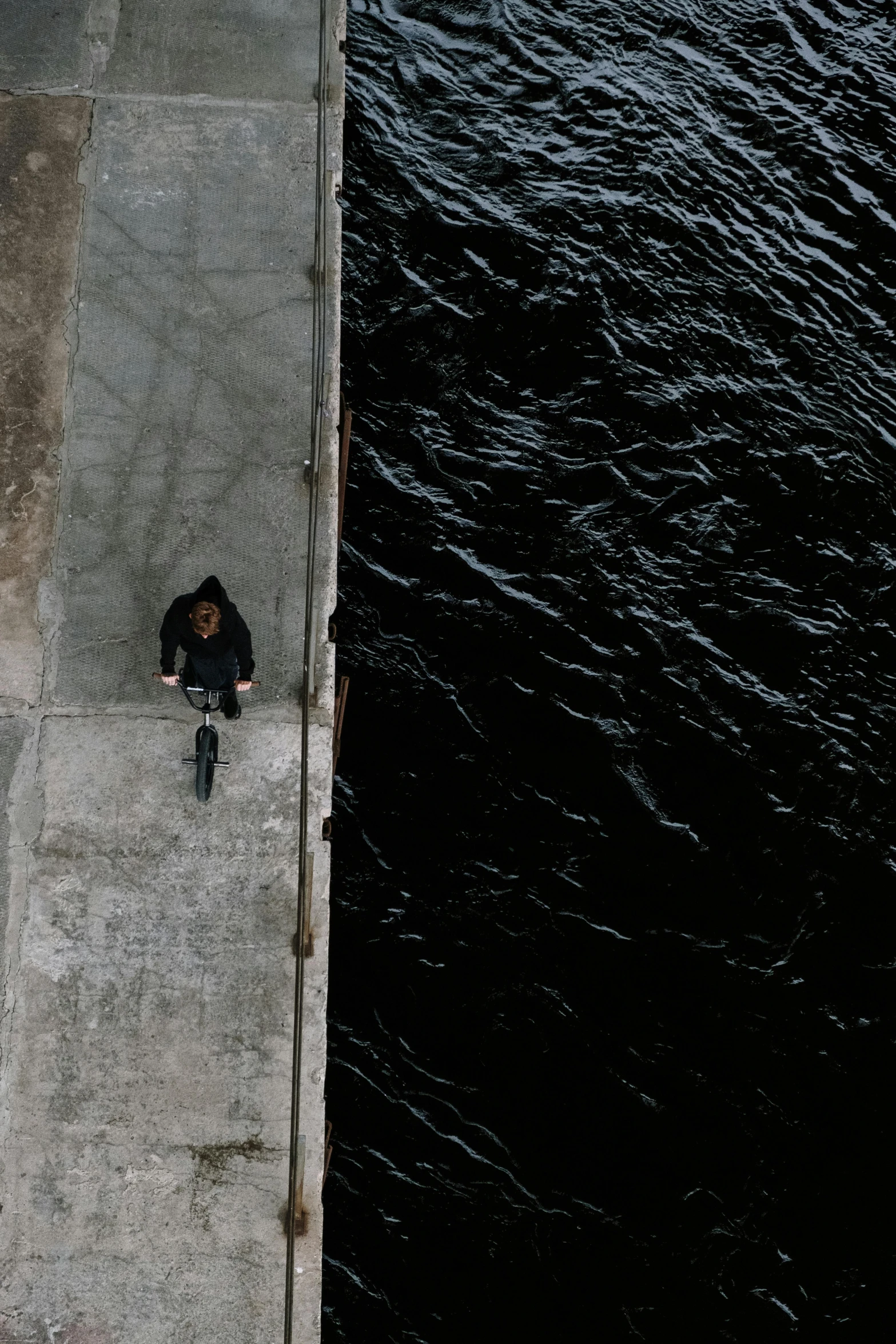 a person sitting on a ledge next to a body of water, inspired by Jan Rustem, pexels contest winner, conceptual art, biker, looking down from above, walking to the right, black