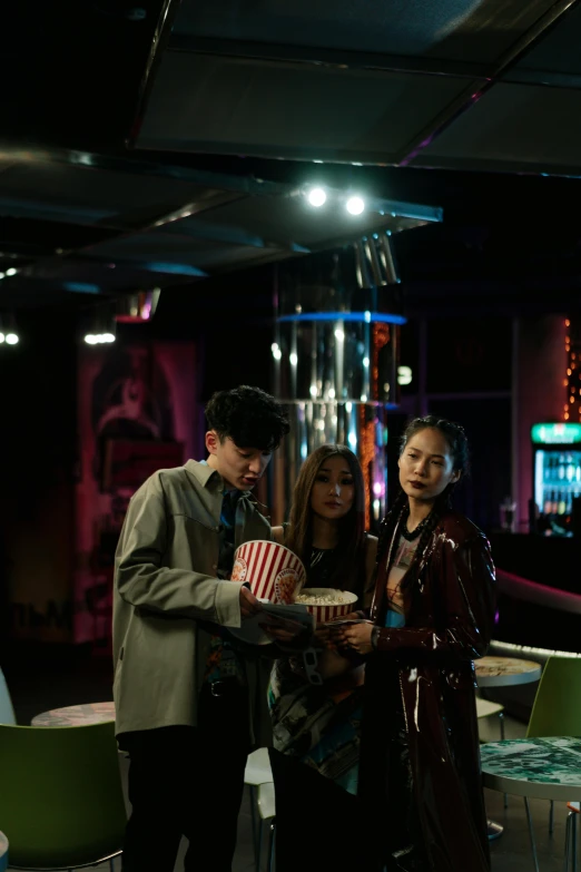 a group of people standing around a table, a hologram, inspired by Nan Goldin, pexels, magical realism, 8 0 s asian neon movie still, [ theatrical ], cold scene, evening lighting