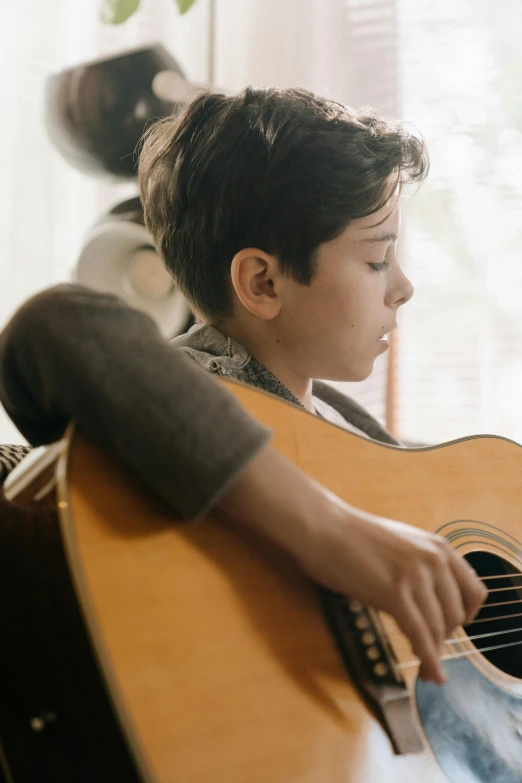 a young boy playing a guitar in a living room, an album cover, pexels contest winner, profile pic, soft natural light, thumbnail, looking her shoulder