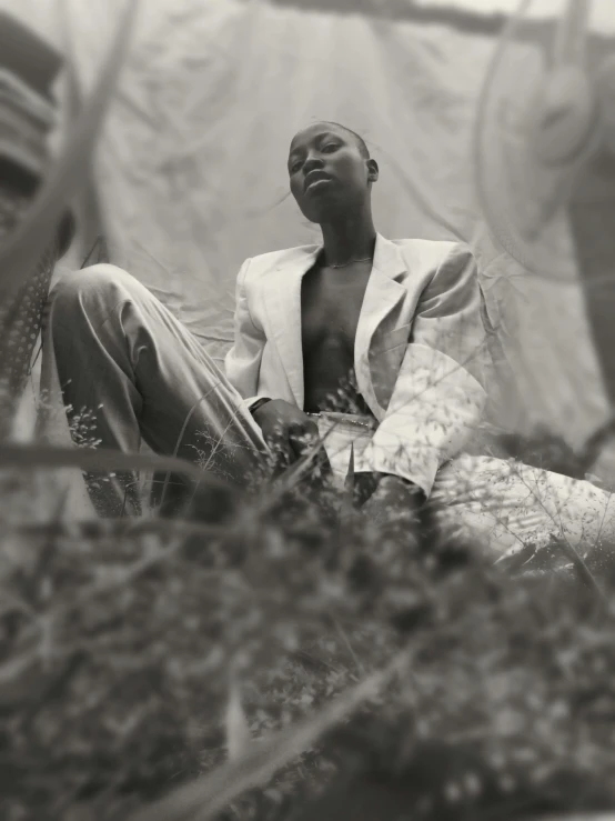 a black and white photo of a man sitting on the ground, an album cover, unsplash, afrofuturism, portrait androgynous girl, white suit, in the high grass, non binary model