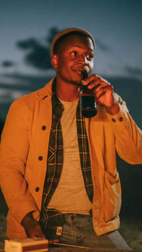 a man standing next to a fire with a drink in his hand, an album cover, by Matt Cavotta, pexels, brown jacket, yellow hue, flannel, dark-skinned