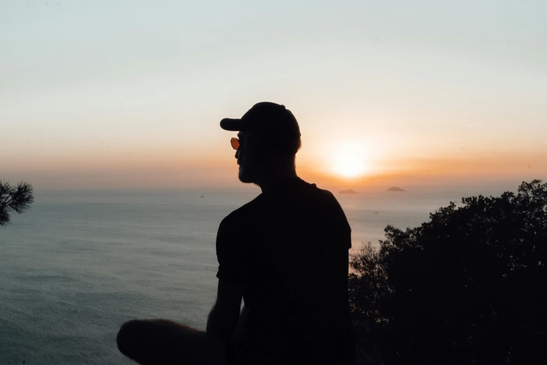 a man sitting on the edge of a cliff watching the sunset, pexels contest winner, wearing sunglasses and a cap, avatar image, profile image, vacation photo