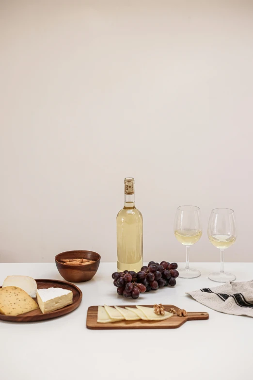 a table topped with plates of food and glasses of wine, on a pale background, white wine bottle, product image, cheeses