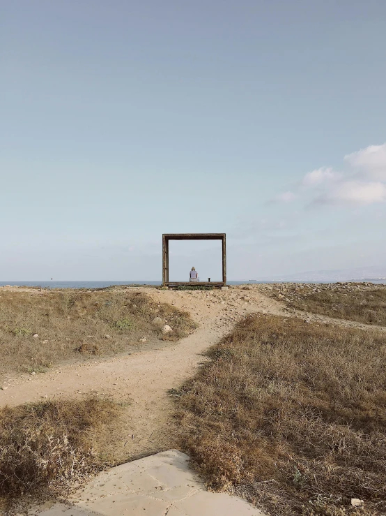 a wooden structure sitting on top of a dry grass covered field, by Attila Meszlenyi, road to the sea, metal framed portal, tiny person watching, cyprus