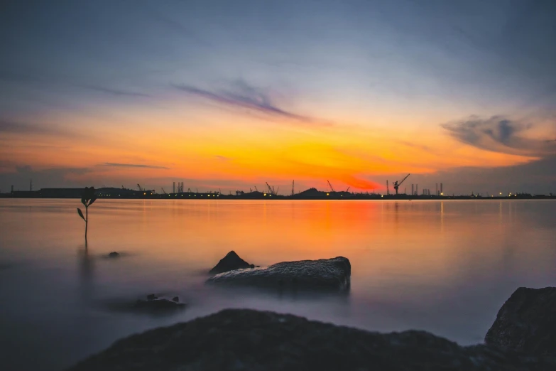 a sunset over a body of water with rocks in the foreground, pexels contest winner, urban surroundings, coloured photo, port scene background, calm environment