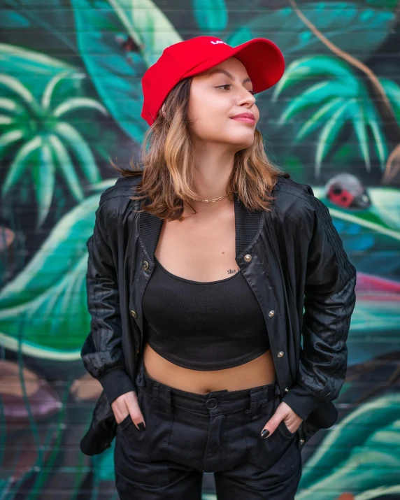 a woman standing in front of a graffiti wall, by Julia Pishtar, wearing a red backwards cap, wearing a cropped black tank top, wearing a fancy black jacket, with palm trees in the back