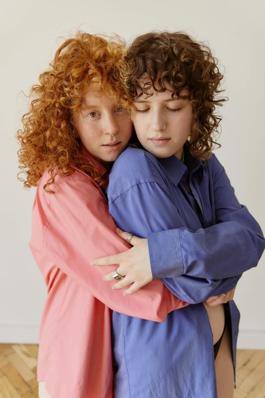 two women hugging each other on a hard wood floor, by Nathalie Rattner, trending on pexels, antipodeans, red haired teen boy, blue and pink shift, long shirt, curls