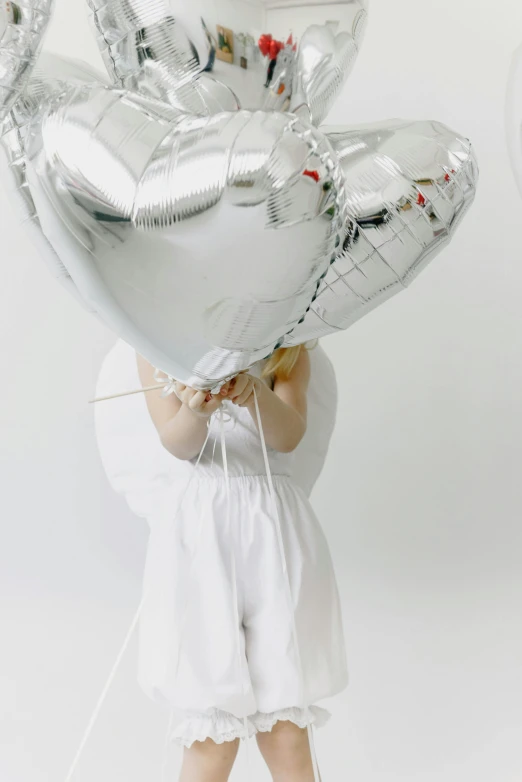 a little girl holding a bunch of silver balloons, wearing white cloths, up close, white space, hearts