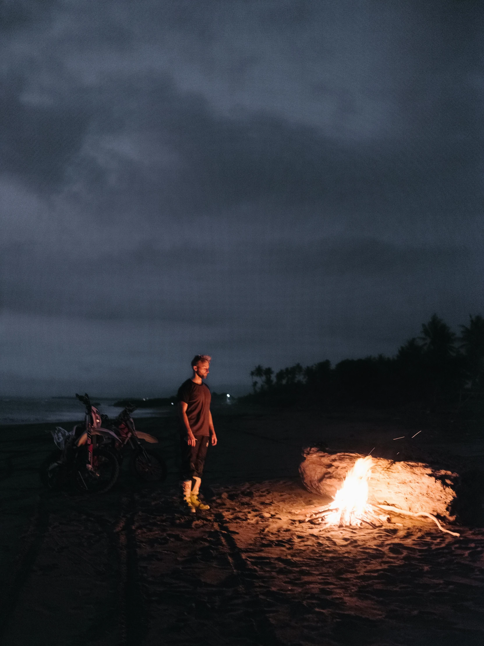 a man standing next to a fire on a beach, by Jessie Algie, overcast lighting, camping, profile image, ultrawide cinematic