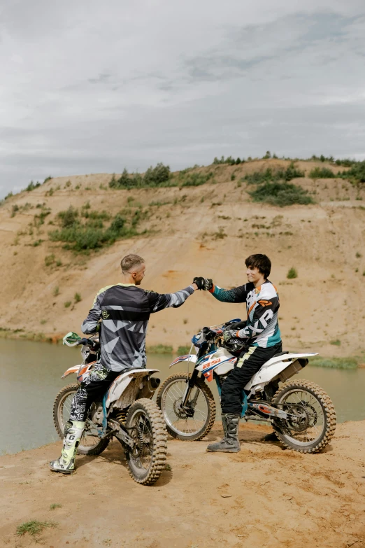 a couple of men standing next to each other on dirt bikes, a picture, unsplash, renaissance, finland, reaching out to each other, 2 0 2 2 photo, river in the background