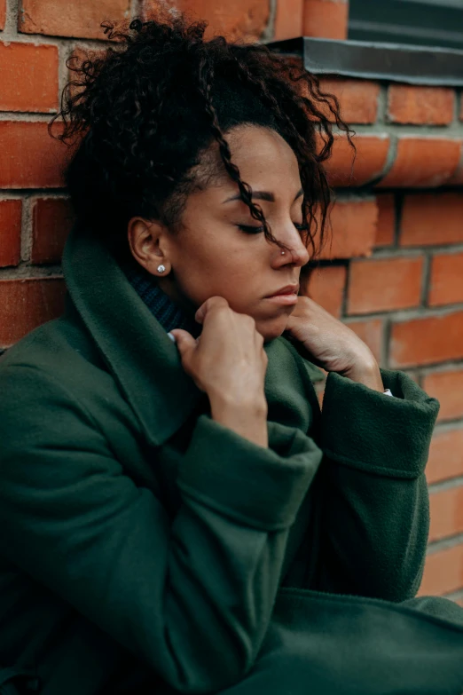 a woman in a green coat leaning against a brick wall, trending on pexels, depressed, mixed race woman, wearing turtleneck, grainy