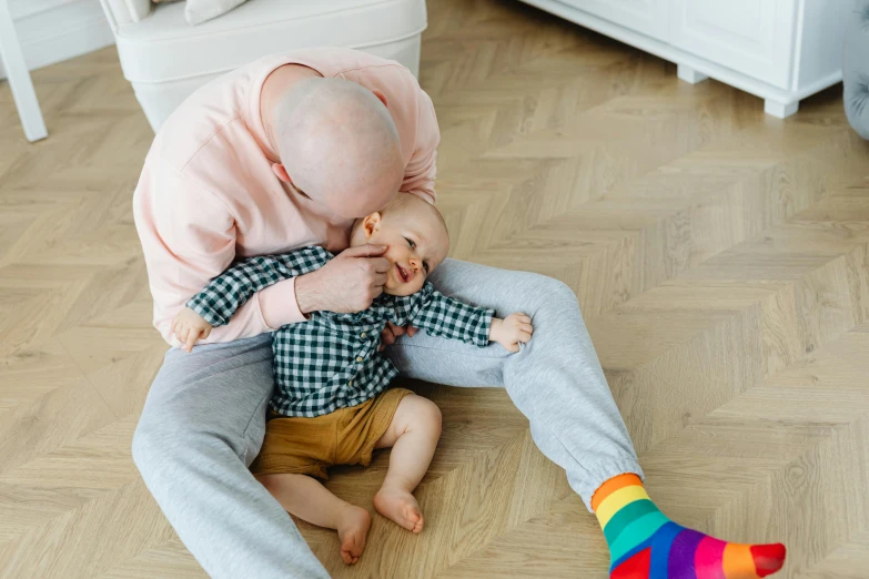 a man sitting on the floor holding a baby, by Emma Andijewska, pexels contest winner, rainbow accents, bald on top, thumbnail, older male