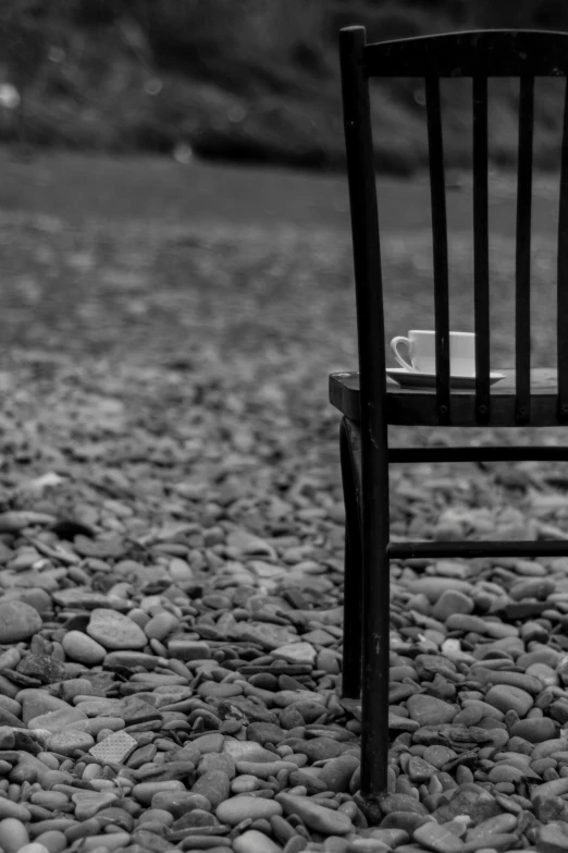 a wooden chair sitting on top of a gravel field, a black and white photo, inspired by Louis Stettner, next to a cup, river, ( ( photograph ) ), tea