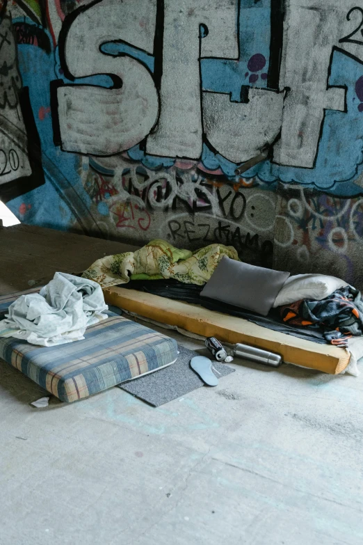 a mattress on the ground in front of a graffiti covered wall, graffiti, two crutches near bench, inside a gang hideout, flat lay, fabrics