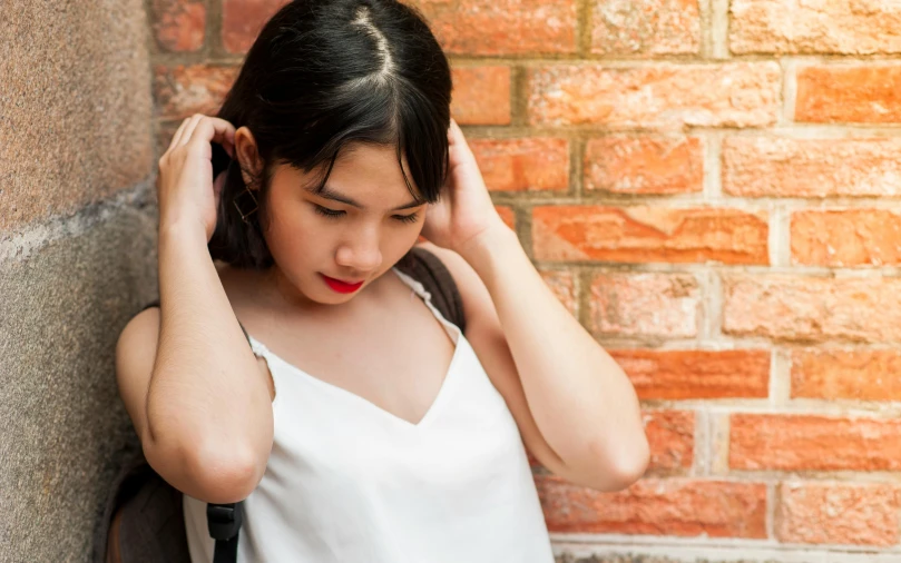 a woman leaning against a brick wall with her hand on her head, inspired by Itō Shinsui, trending on pexels, wearing white camisole, portrait of depressed teen, tinnitus, vietnamese woman