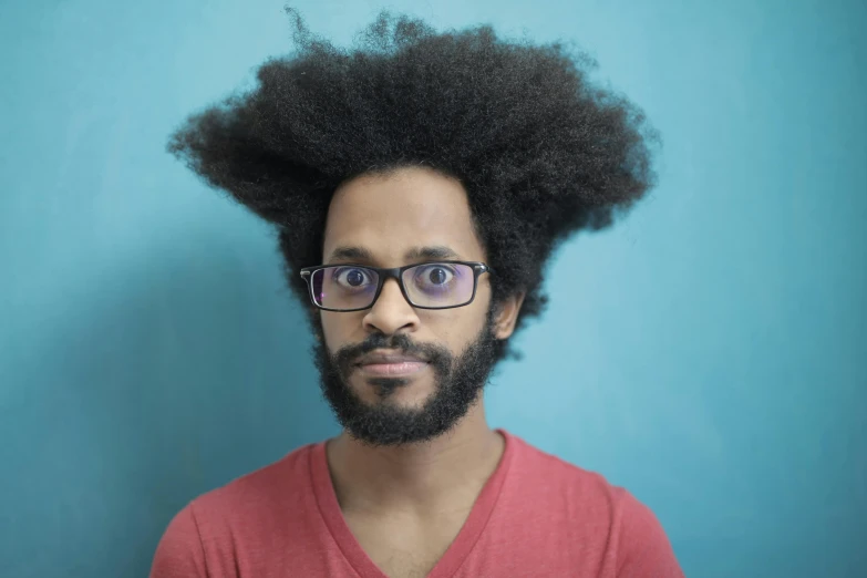 a man with a beard and glasses standing in front of a blue wall, an album cover, by Washington Allston, pexels contest winner, fantastic realism, afro hair, crazy hair, on grey background, edu souza