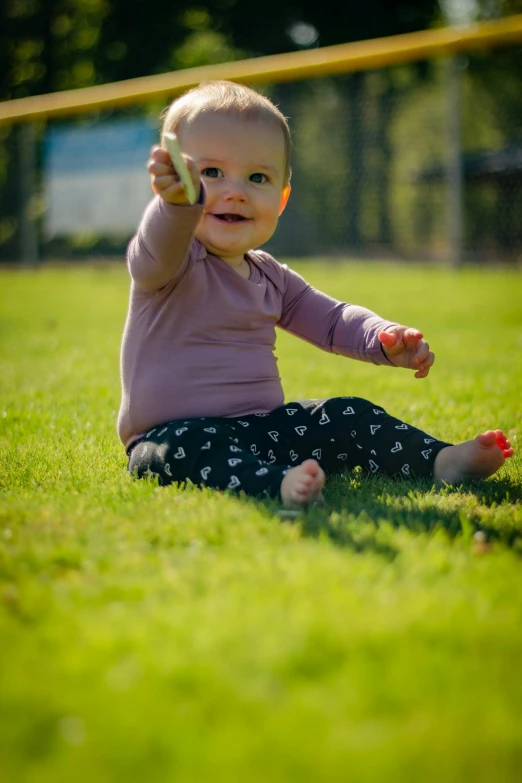 a baby sitting on top of a lush green field, pointing at the camera, stretching her legs on the grass, colour photo, square