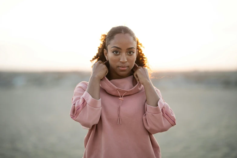 a woman standing on top of a sandy beach, a portrait, trending on pexels, happening, wearing a pastel pink hoodie, light-brown skin, wearing turtleneck, vanessa morgan