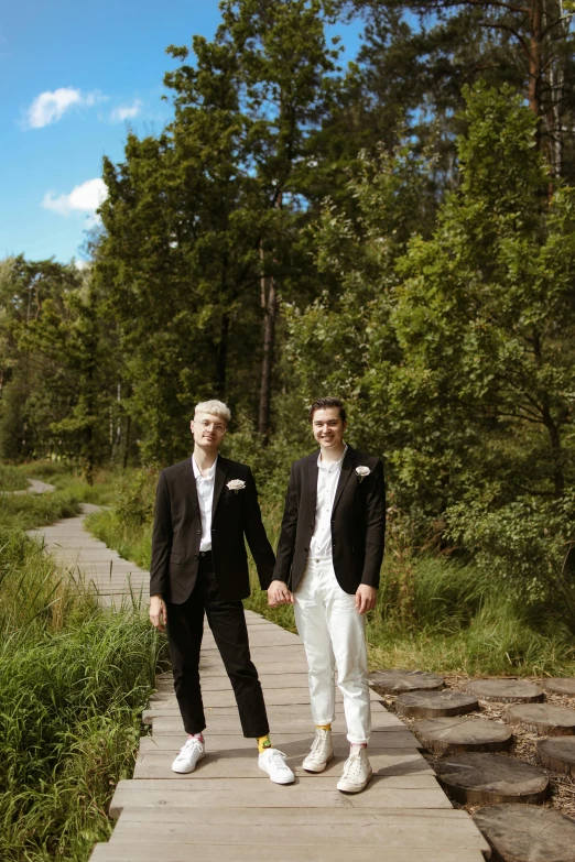 a couple of men standing on top of a wooden walkway, an album cover, by Anato Finnstark, wearing a white tuxedo, on forest path, holding each other hands, non-binary