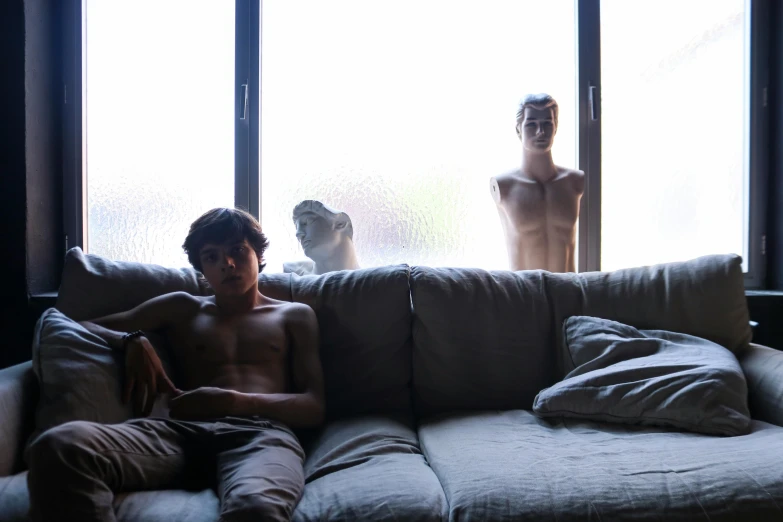 a man sitting on top of a couch next to a window, a statue, by Elsa Bleda, pexels contest winner, hyperrealism, as 3 figures, teenage boy, shirtless, soft and blurry