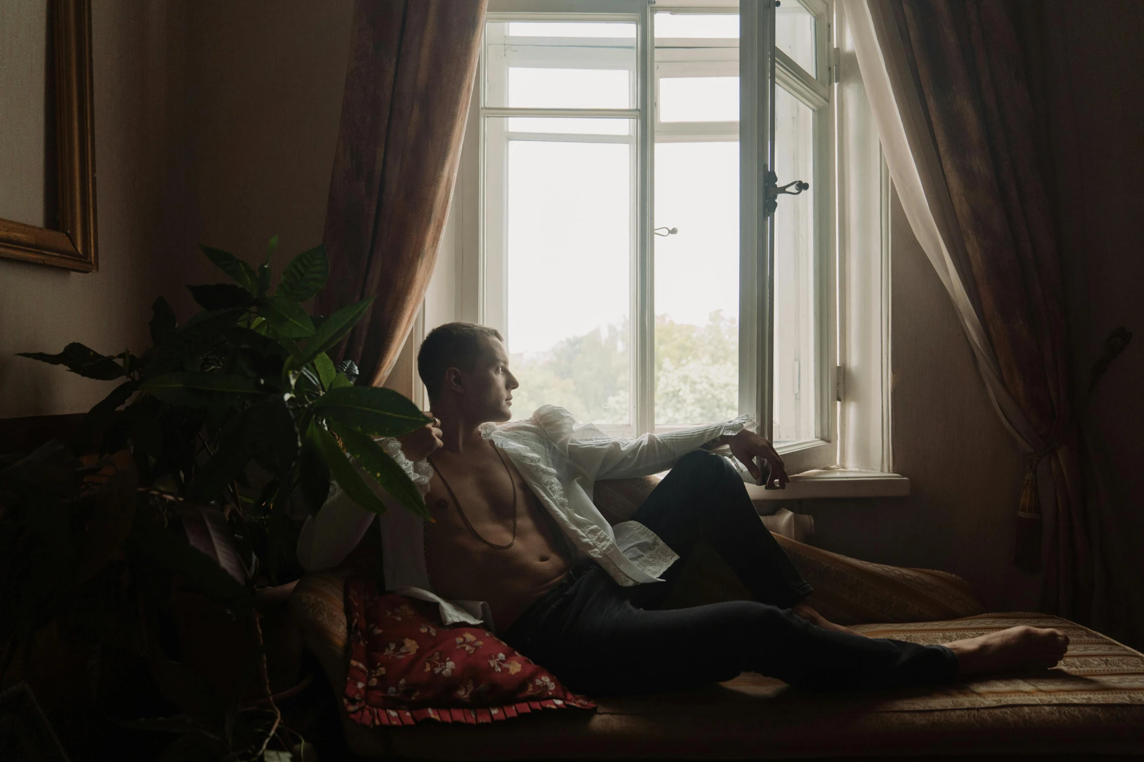 a man sitting on top of a bed next to a window, inspired by Elsa Bleda, vitalik buterin, relaxing on the couch, yulia nevskaya, lgbtq