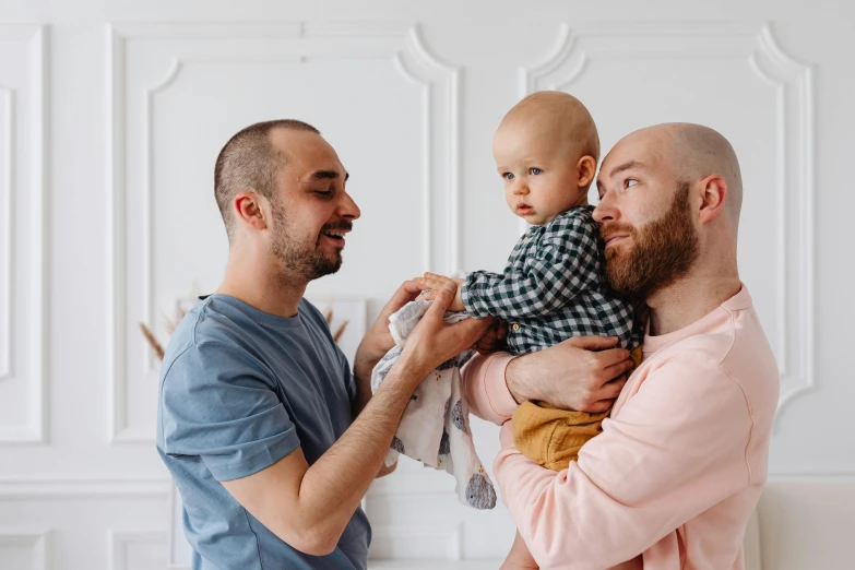 a man holding a baby while standing next to another man, pexels contest winner, antipodeans, brunette boy and redhead boy, bearded, bald, nursing