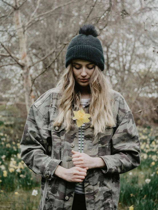 a woman standing in a field of daffodils, unsplash, wearing beanie, camouflage uniform, holding a flower, wearing a black jacket