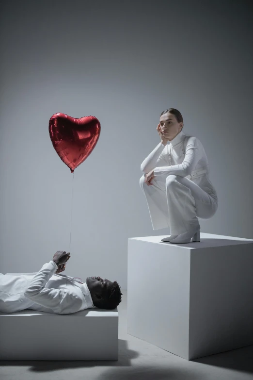 a man sitting on top of a white box next to a woman, a surrealist sculpture, inspired by Marina Abramović, pexels contest winner, red hearts, balloon, frank dillane as puck, showstudio