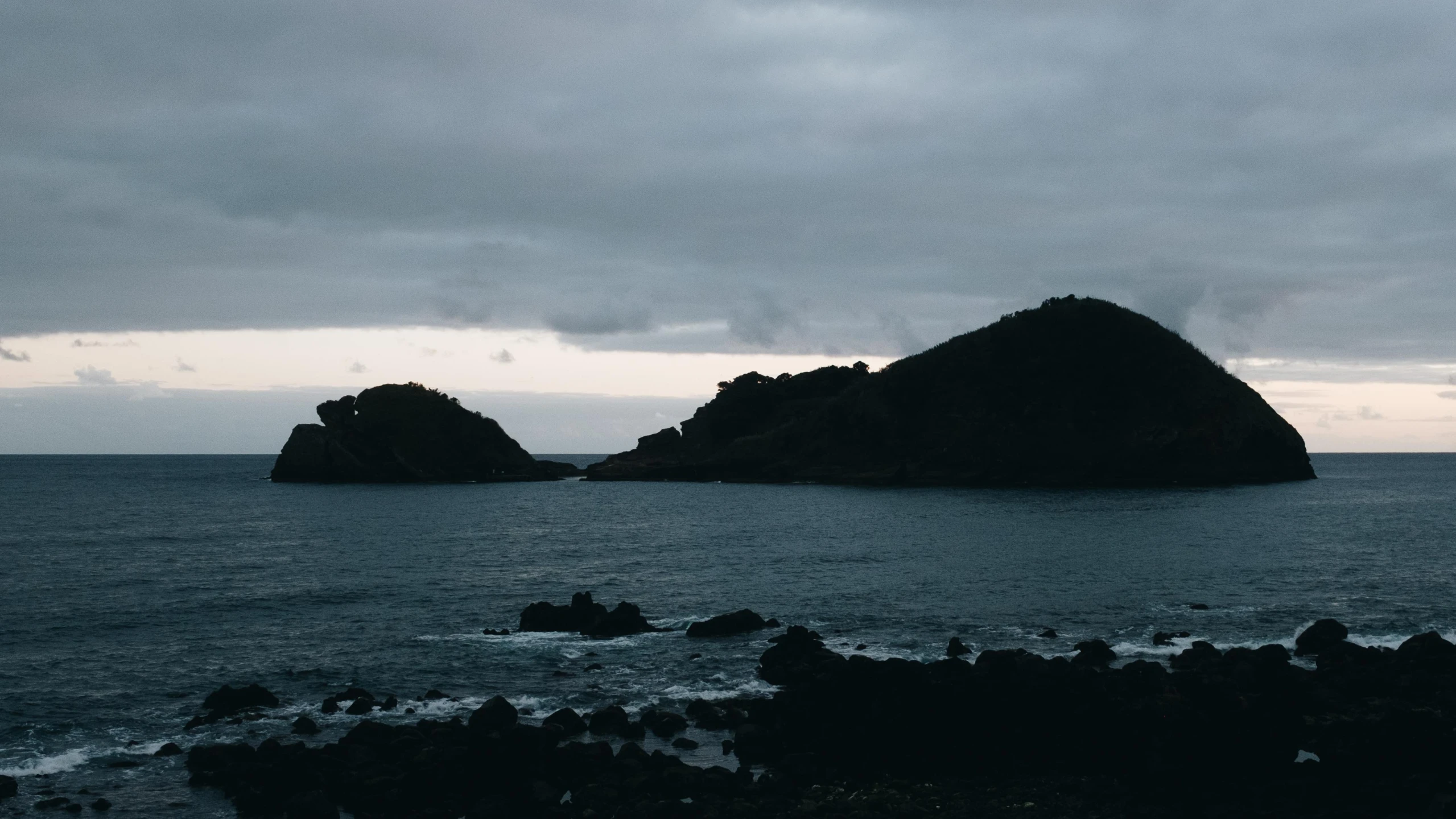 a body of water surrounded by rocks under a cloudy sky, unsplash, australian tonalism, two medium sized islands, wellington, low quality photo, early evening