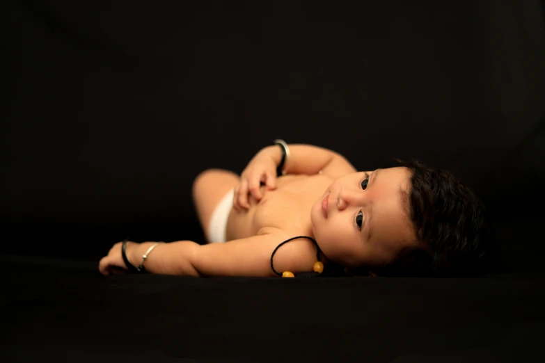 a close up of a baby laying on a black surface, by irakli nadar, unsplash, art photography, black jewellery, full body picture, vinayak, looking to his side