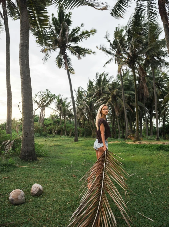 a woman standing on top of a lush green field, by Jessie Algie, land art, coconut palms, holding a 🛡 and an 🪓, near the beach, holding a wooden staff