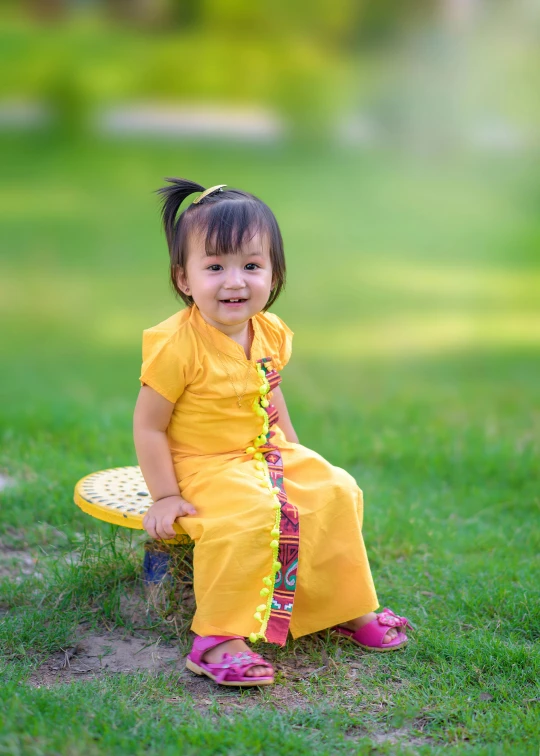 a little girl sitting on top of a yellow bench, inspired by Tang Di, pixabay contest winner, happening, wearing authentic attire, sitting on green grass, full - length photo, small chin