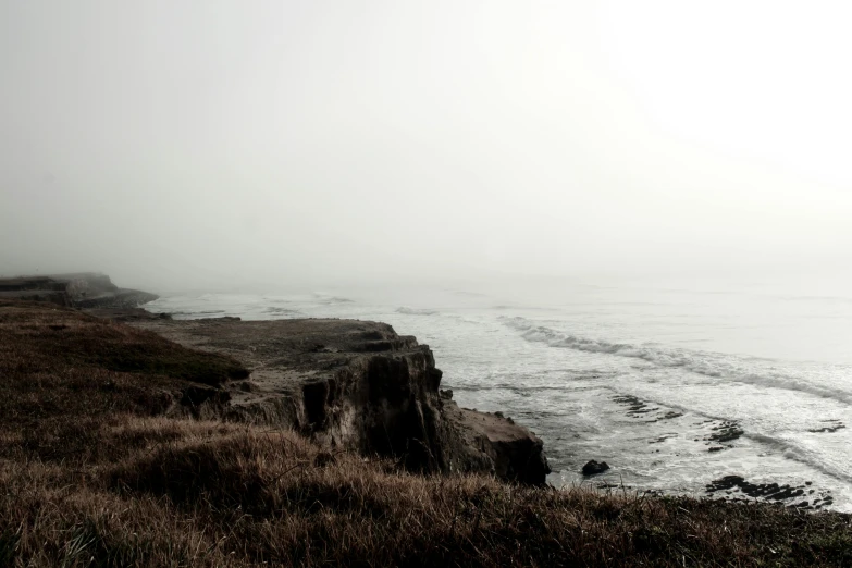 a man standing on top of a cliff next to the ocean, a black and white photo, unsplash, romanticism, brown mist, background image, photography of todd hido, moist foggy