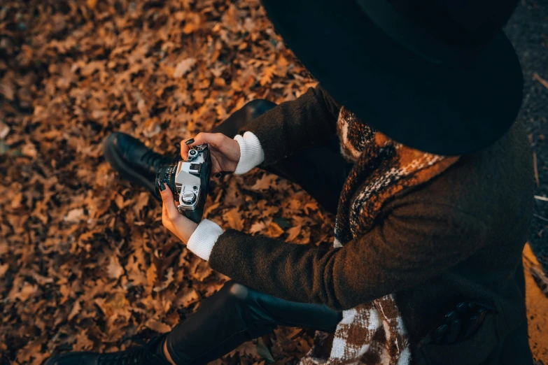 a person sitting on the ground holding a camera, by Emma Andijewska, pexels contest winner, 🍂 cute, holding a can of beer, black stetson and coat, holding controller