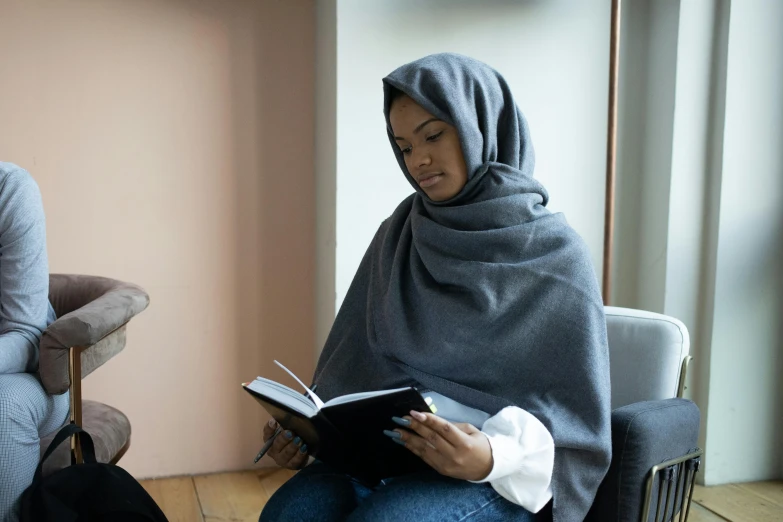 a woman sitting in a chair reading a book, hurufiyya, wearing a grey hooded sweatshirt, wrapped in a black scarf, riyahd cassiem, holding notebook