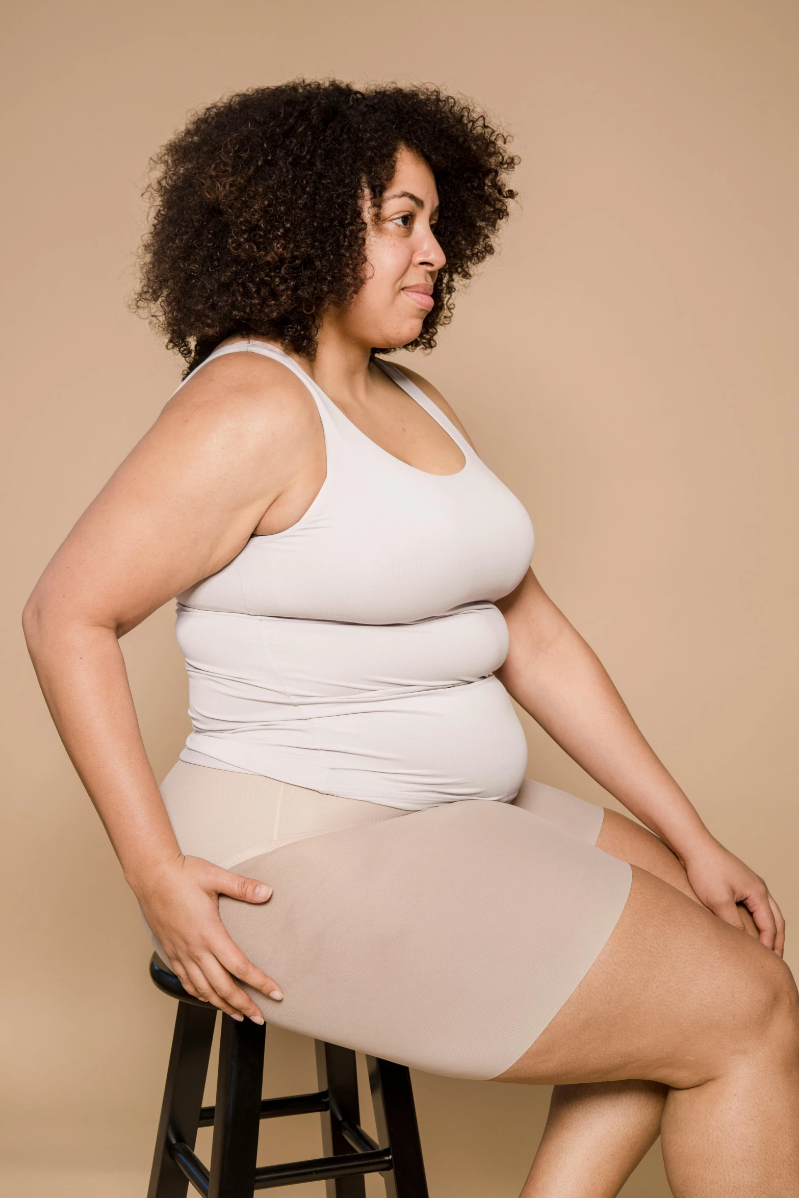 a woman sitting on top of a stool, half - body composition, wearing a tanktop and skirt, big stomach, taupe