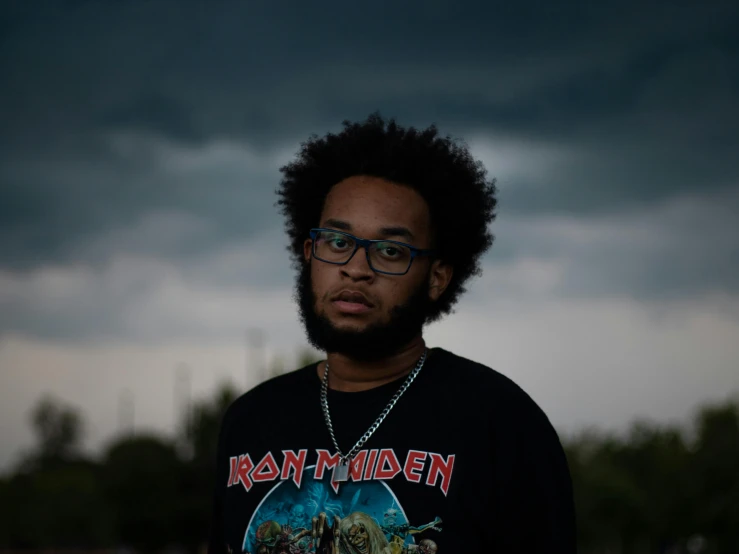 a man with glasses standing in front of a cloudy sky, an album cover, by Stokely Webster, pexels contest winner, realism, light skin, (night), dark brandon, shot on 70mm