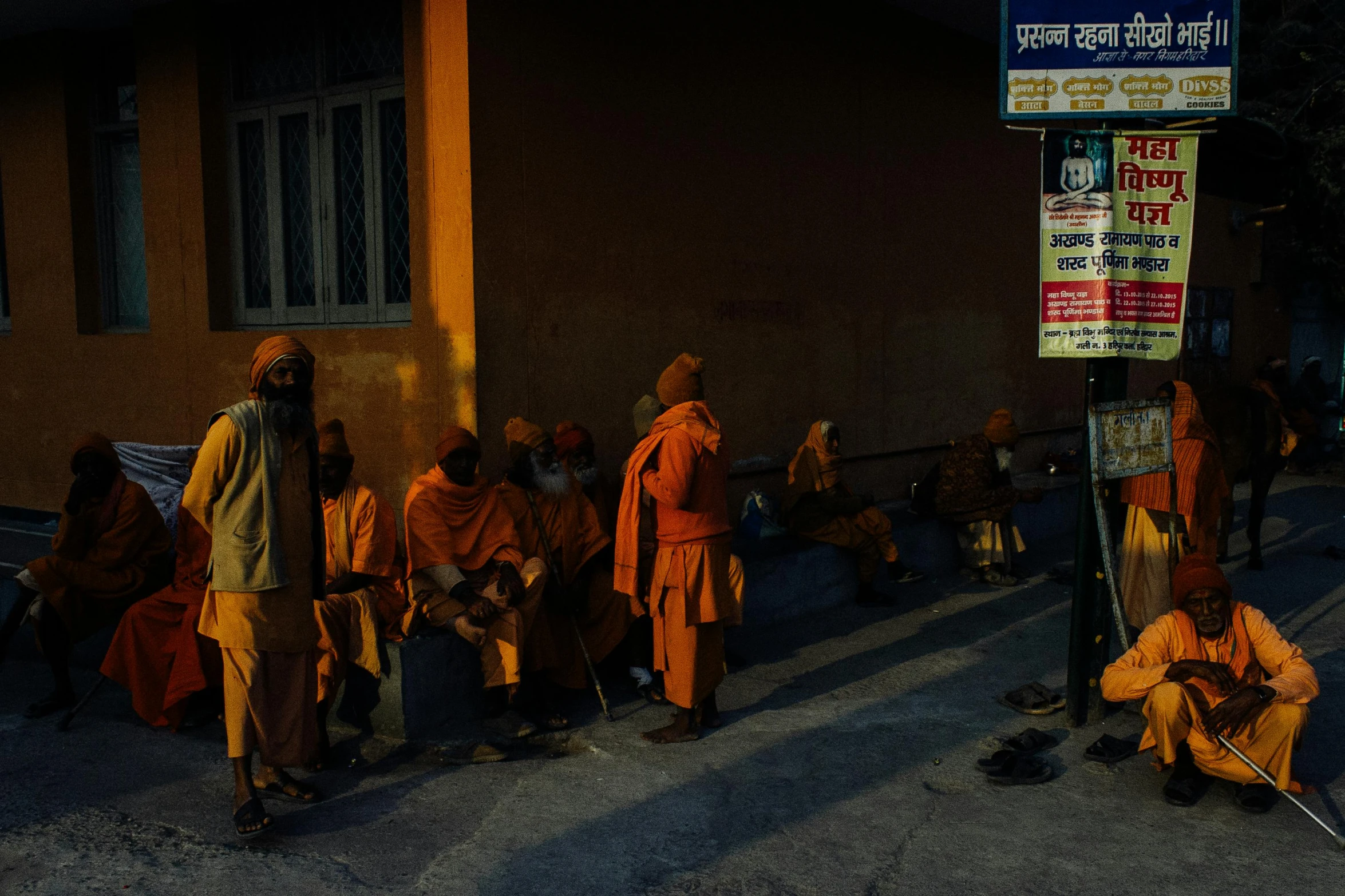 a group of people sitting on the side of a road, pexels contest winner, bengal school of art, orange yellow ethereal, train station, holy man looking at ground, dark city bus stop