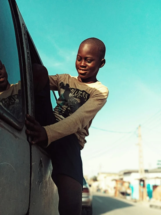 a young boy leaning on the side of a car, by Nathalie Rattner, pexels contest winner, happening, african aaron paul, slide show, ignant, at full stride