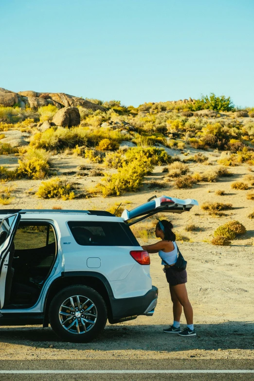 a woman standing next to a car with a surfboard on top of it, rocky terrain, serene desert setting, campsites, multiple stories