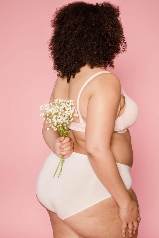 a woman in a white underwear holding a flower, pastel pink skin tone, plus-sized, her back is to us, wearing bra